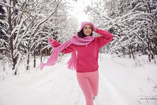 woman run on a path in the woods in pink clothes a jacket a knitted scarf and a hat stands in a snowy forest in winter