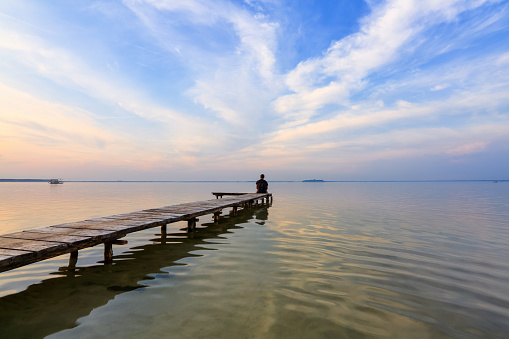Man seated on a wooden jetty, pier looking a blue sky with cloud, silhouette reflected on the calm water. Beautiful lake. Summer vacation. Free space for text.