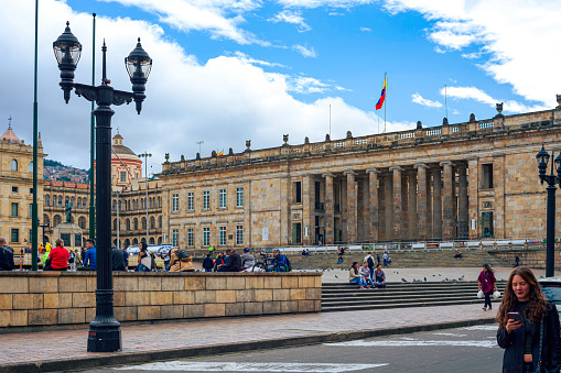 Bogota, Colombia - January 27, 2017: Looking across Plaza Bolivar, in the Andean capital city of Bogota in South America, to the building that is the official place of business for both houses of parliament: the Senate and the Congress. Completed in 1926, it was designed in the 19th Century by the Danish Architect Thomas Reed. Local Colombian people nonchalantly walk past the building of prime importance to the Country, as they go about their daily lives. The Colombian national flag flies over the building. Photo shot in the afternoon sunlight; horizontal format. Copy space.