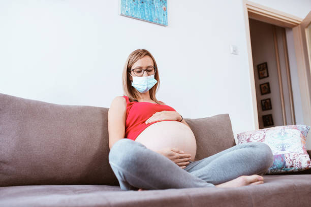 istock Young pregnant woman with a protective mask on her face is sitting on a sofa 1298020657