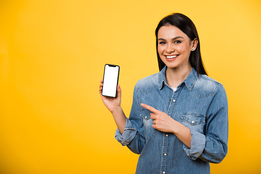 Happy caucasian brunette girl in blue casual shirt, shows finger at phone with blank white screen, looks at camera and smiling with friendly smile, standing against isolated orange background
