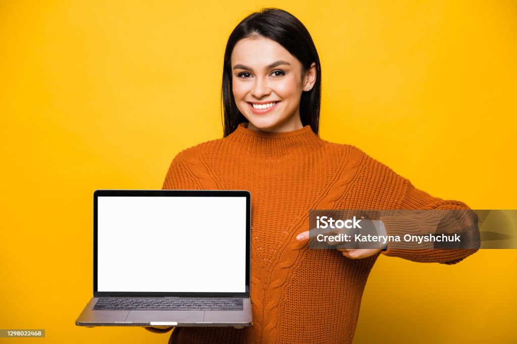 Young smiling caucasian brunette woman dressed in orange sweater, holds laptop with blank white screen, shows finger at  it, looks at camera and friendly smiling, standing on isolated orange background. Copy space Laptop Stock Photo