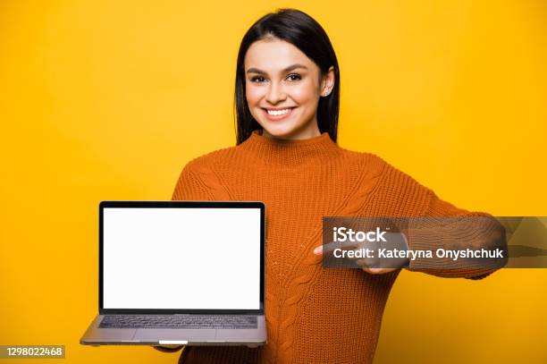 Young Smiling Caucasian Brunette Woman Dressed In Orange Sweater Holds Laptop With Blank White Screen Shows Finger At It Looks At Camera And Friendly Smiling Standing On Isolated Orange Background Copy Space Stock Photo - Download Image Now