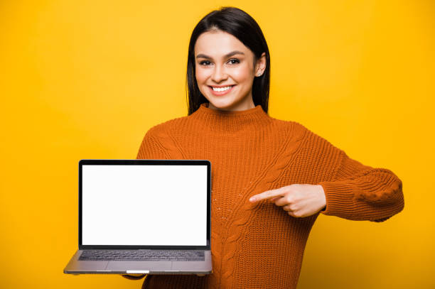 jovem morena branca sorridente vestida com suéter laranja, segura laptop com tela branca em branco, mostra o dedo para ele, olha para a câmera e sorria amigável, de pé sobre fundo laranja isolado. copiar espaço - teenage girls cheerful smiling one person - fotografias e filmes do acervo