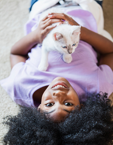 A 9 year old African-American girl lying on the floor, holding a kitten. The girl is smiling, looking at the camera.