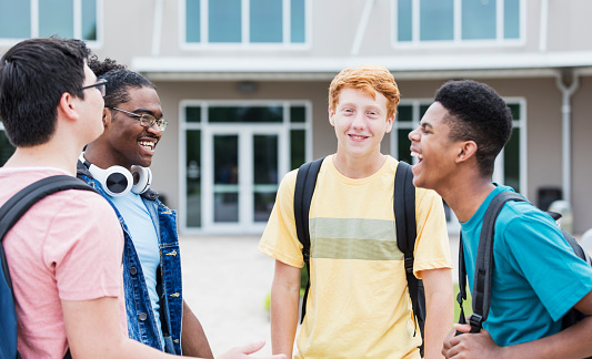 A multi-ethnic group of four high school students, teenage boys 15 to 17 years old, standing in front of the school building, conversing. They are a diverse group, including two African-Americans, a mixed race Caucasian/Asian and a redhead.