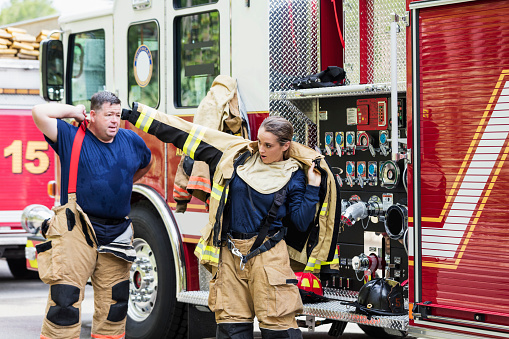 Two firefighters getting ready to answer a call, putting on their fire protection suits, standing next to a fire engine. One of them is a young woman in her 20s. Her partner is a mature man in his 40s.