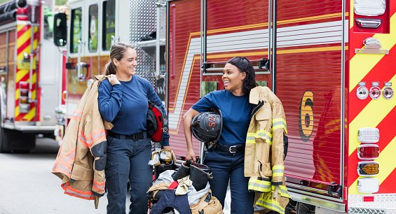 Two multi-ethnic female firefighters carrying their equipment and fire protection suits, conversing as they walk by two fire engines.