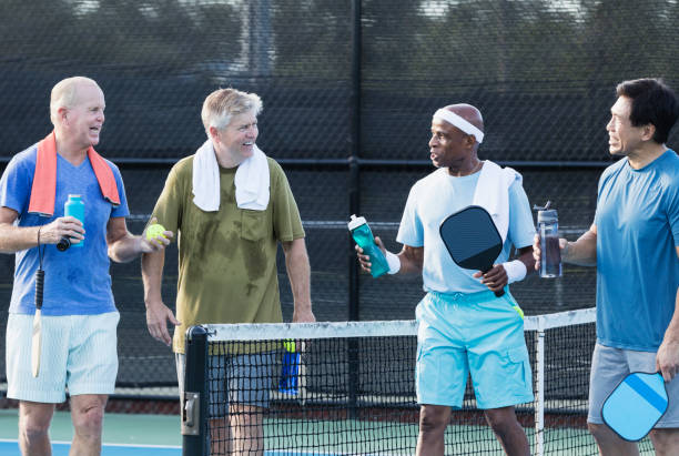 Group of senior men on pickleball court, taking break A multi-ethnic group of four mature and senior men walking off a pickleball court, smiling and conversing. They are taking a break after playing, towels around their shoulders, holding water bottles. The African-American man is the oldest, in his 70s. The others are in their 50s and 60s. paddle ball stock pictures, royalty-free photos & images