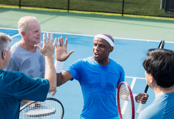 Men playing tennis, high-five A multi-ethnic group of four mature and senior men playing tennis outdoors. They are standing together at the net, smiling. The African-American player, a senior man in his 70s, is giving a high-five to one of his opponents. tennis senior adult adult mature adult stock pictures, royalty-free photos & images