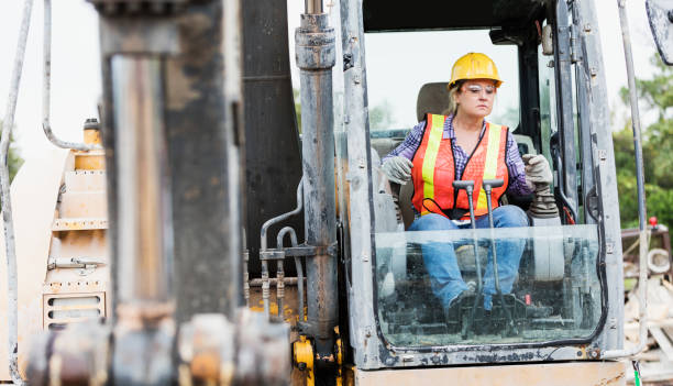 Hispanic female construction worker driving earth mover A mature Hispanic woman in her 40s working at a construction site, operating an earth mover. She is wearing a hardhat, reflective vest, and safety goggles. construction worker stock pictures, royalty-free photos & images
