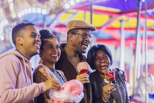 A senior African-American couple in their 60s with their adult grandchildren, in their 20s, having fun at a traveling carnival, eating cotton candy and candy apples.