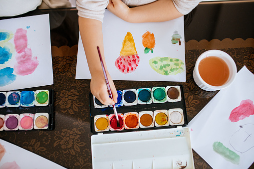 Two schoolgirls sitting at table, painting with water colors and having fun. Focus on the painting on the right