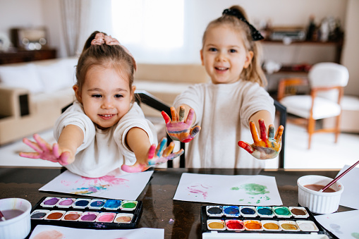 Two schoolgirls sitting at table, painting with water colors and having fun. Focus on the painting on the right