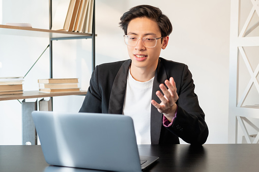 Online presentation. Business webinar. Internet communication. Confident Chinese male CEO in glasses discussing project with team using laptop at light workplace.