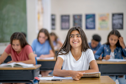 A high school girl of Indian ethnicity smiles widely at the camera as she is sitting at her desk in her classroom.