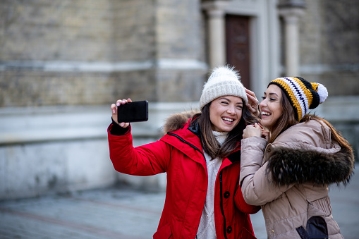 Two girls in knit hats are taking selfies, smiling and enjoying in their tour downtown. They haven't seen each other for too long and they are happy to spend some quality time together outdoors, on city streets, during winter.