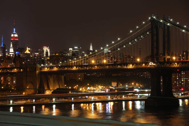 A stunning nighttime view capturing the lit Brooklyn Bridge, with cityscape elements and reflections creating a harmonic urban ambiance. The photograph highlights an iconic landmark and dynamic aesthetics of New York City.
