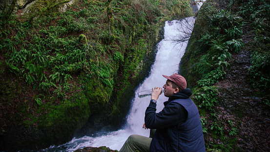Hiker enjoying the Columbia River Gorge in Oregon by watching a waterfall, he has a cold water bottle next to him.