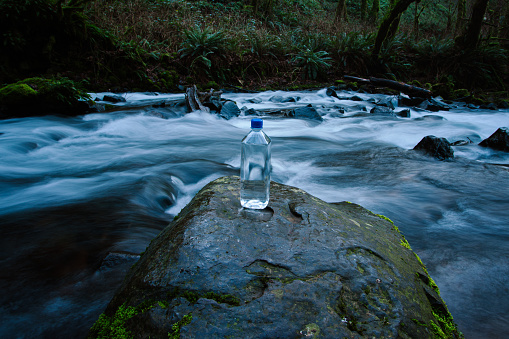 Stand alone water bottle outdoors in a nature scene. Placed there by a hiker. Focus is on the water bottle with a flowing stream of water in the background.