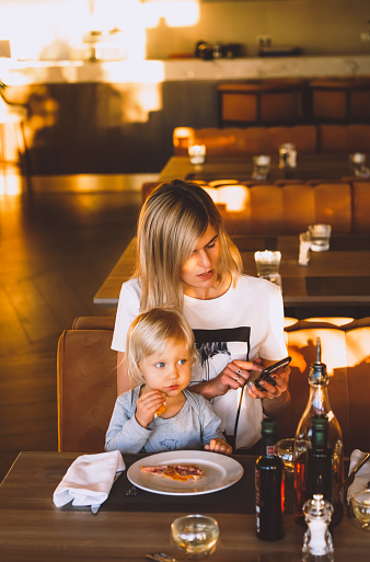 Young blond mother relaxing in sunny indoor cafe text messaging with her phone with her little cute child eating pizza.