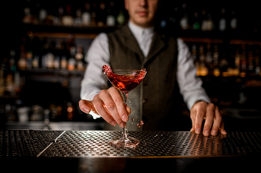 beautiful glass with splashing drink is placed on metal surface of the bar and hand of man bartender holds it