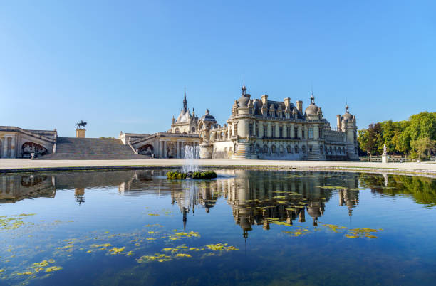 Chantilly, France - September 21 2020: View of the Chateau de Chantilly from Le Notre Garden with a fountain in the foreground and reflection of the castle in a pond - France