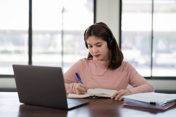 istock Young Asian girl student wears wireless headphones write on the notebook to study language online watch and listen to the lecturer, webinar via video call e-learning at home, distance education 1298085240