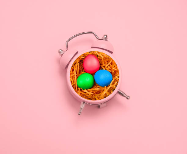 Top view creative minimal art collage alarm clock and bird nest with painted eggs on a pastel pink background. Copy space. Easter minimal card.