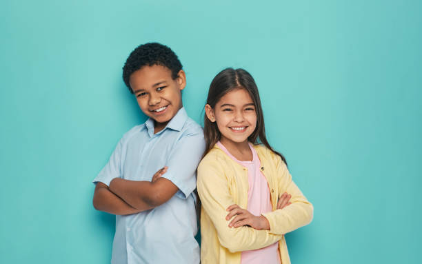 chica asiática y niño afroamericano de pie espalda con los brazos cruzados sobre un fondo turquesa - cheerful happiness smiling teenager fotografías e imágenes de stock