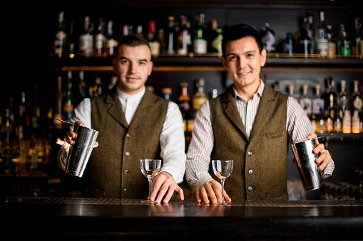 two smiling male bartenders with shaker cups in their hands stand behind the bar counter