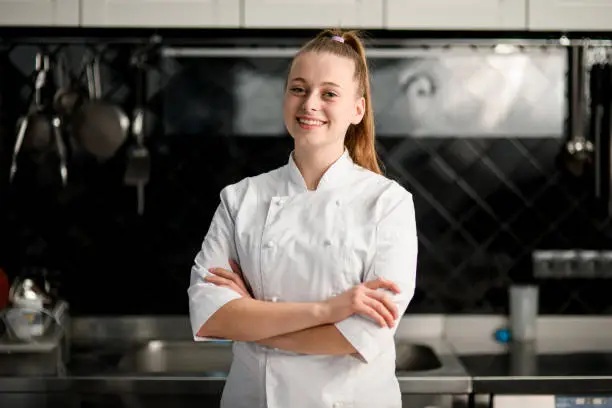 young beautiful smiling woman chef with arms crossed wearing in white suit at kitchen. Blurred background