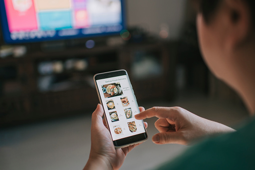 Online food delivery mobile app shown on smart phone screen hold by asian woman hands in living room
