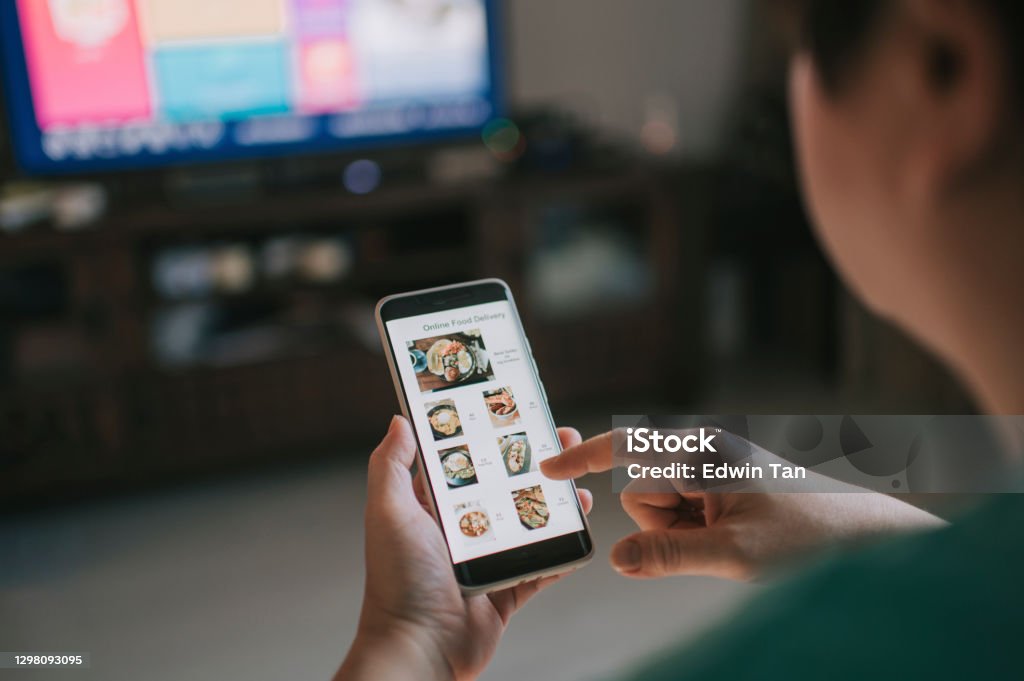 Online food delivery mobile app shown on smart phone screen hold by asian woman hands in living room Online food delivery mobile app shown on smart phone screen hold by asian woman hands indoor Television Set Stock Photo
