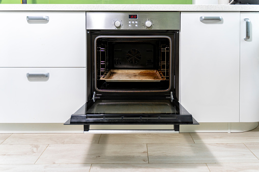 the door of a dirty oven is open in a bright kitchen.