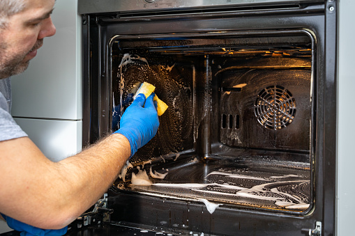 Caucasian big man washes the oven with foam and sponge. House cleaning