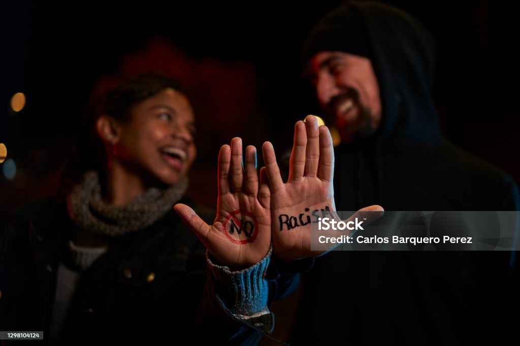 A young white man and a young black woman smiling, showing a handwritten message against racism. Anti-racism Stock Photo