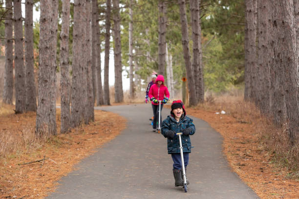 Children riding their scooters on a small hill in the woods. Taken on a late fall/early winter day. The trail is paved, and there are tall pine trees all around them.