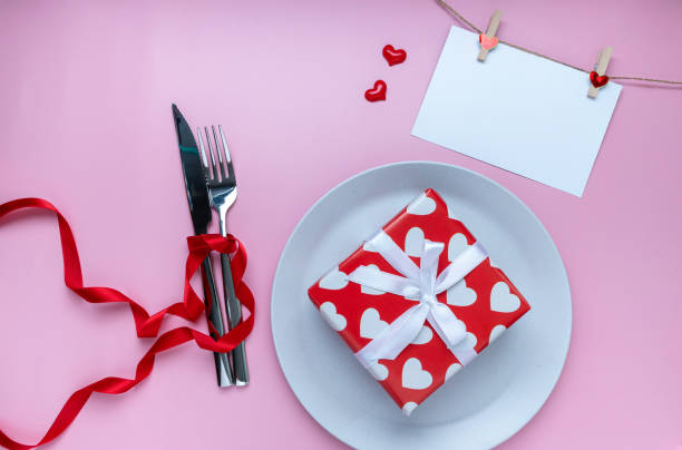 istock Romantic dinner table. Gift or present box, greeting card, mini heart, plate, fork and knives with ribbon on pink background. for banner. The concept of Valentine Day. top view, copy space 1298113742