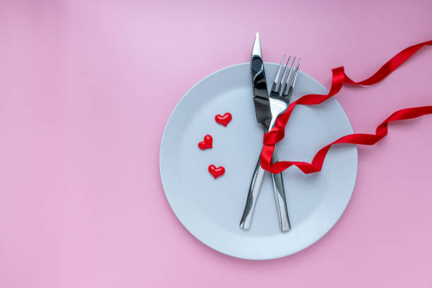 istock Romantic dinner table. plate, fork and knives with ribbon on pink background. for banner. The concept of Valentine Day. top view, copy space 1298113833