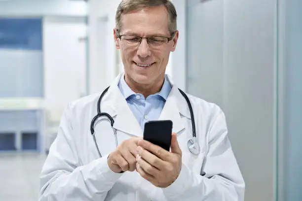 Photo of Old male doctor holding smartphone using mobile phone e health technology in hospital. Modern tech medical healthcare patients virtual consultation app, telehealth online, tele medicine appointment.