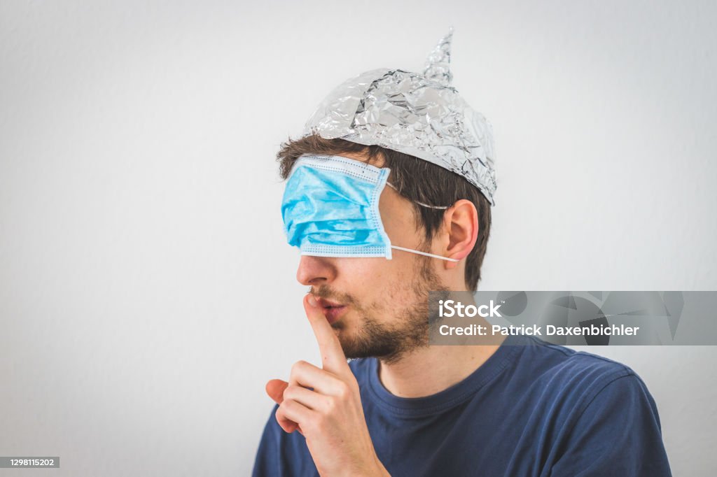 Conspiracy theory concept: Young man with face mask over the eyes and aluminum hat is making a psst! gesture Young angry man with face mask over the eyes and aluminum hat is doing a psst! gesture QAnon Stock Photo
