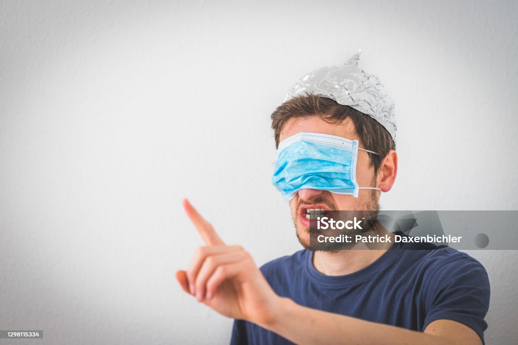 Conspiracy theory concept: Young angry man wearing aluminum hat and face mask over the eyes is gesturing angry. Young angry man with aluminum cap and face mask over the eyes is gesturing angry. Conspiracy theory concept. QAnon Stock Photo