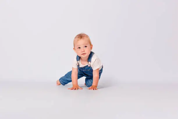 Photo of Cute year-old baby girl in a blue denim jumpsuit sits on white background. a child's photo shoot