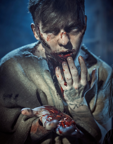 Young man with dull eyes dressed and made up like a zombie. He is looking at the camera. His mouth is filled blood. Zombie man is holding worms in his hands. Studio shooting, portrait, waist-length