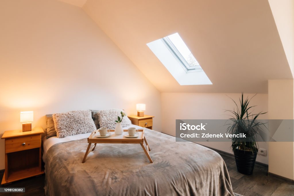 Small attic bedroom with morning breakfast service Small attic bedroom in a family house with morning breakfast service on the bed. The bed also has a fluffy blanket and two fluffy pillows, on the sides there are bedside tables with lit lamps. Family Stock Photo
