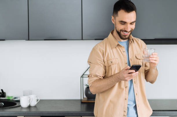 istock Bearded caucasian man use smartphone, chatting with friends or browsing internet. Happy busy man hold a glass of pure water, follow healthy lifestyle and drink the daily rate of water 1298133450