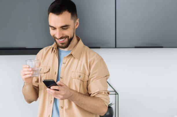 istock Bearded caucasian man use smartphone, chatting with friends or browsing internet. Happy busy man hold a glass of pure water, follow healthy lifestyle and drink the daily rate of water 1298133452