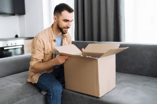 istock Home delivery. Stylish caucasian guy got his order, unpack a parcel. Happy man with interest look inside cardboard box, sitting on sofa, the guy shopping in internet stores, buying new clothes online 1298133455