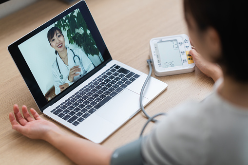 A woman is measuring her blood pressure while consulting with a doctor via telemedicine video call on a laptop at home